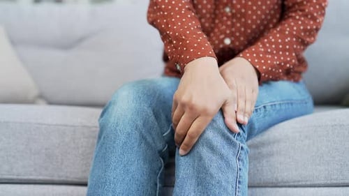 Close up. Female hands massaging knee leg muscles while sitting on sofa in living room at home.