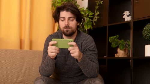 Adult Man Sitting on the Sofa in the Room Playing Video Game on Smartphone Watching Media Close Up