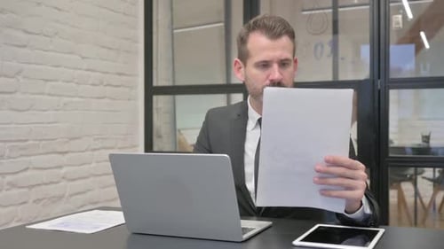 Man Reviews Documents at Desk with Laptop