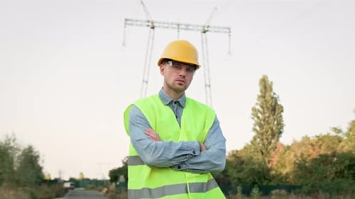 Serious Power Line Inspector Posing for Camera Against Transmission Tower