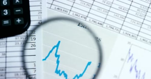 A hand with a magnifying glass passes over a financial newspaper and financial bag with a business