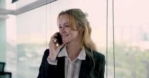 Caucasian female adult businesswoman smiling while talking on smartphone near window in office