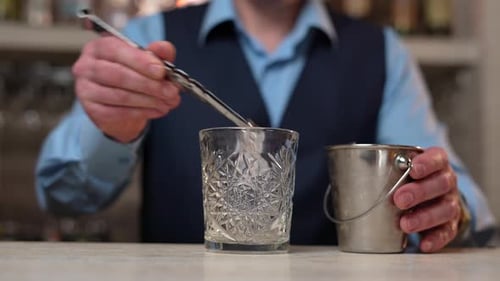 Bartender with steel tongs Puts Ice in Whisky Glass. Close up