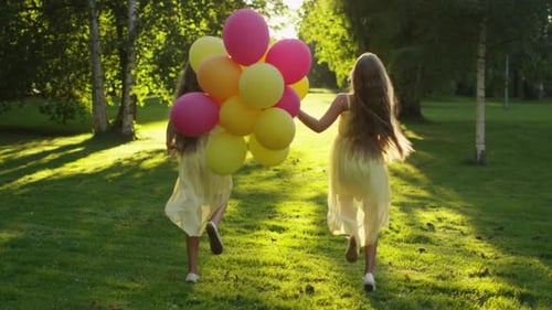 Twin Girls is Running at Park with Balloons in Hands at Sunset Time