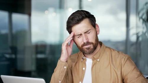 Businessman suffering from headache while working on laptop sitting at the workplace in office.