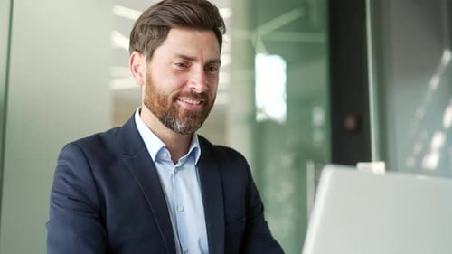 Smiling businessman in a formal suit typing on a laptop sitting at a workplace in business office.