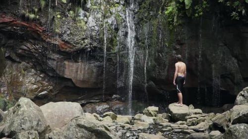 Panning up to man on rock gazing at small waterfall in flourishing jungle