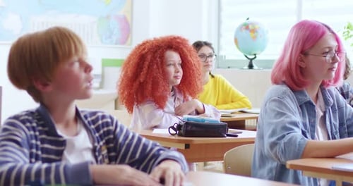Cute Small Clever Schoolgirl with Red Curly Hair Sitting in Classroom Among Other Teens Pupils and