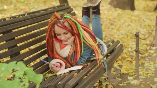 Young Woman with Dreadlocks Drawing Writing in Notepad Lying on Park Bench in Golden Autumn