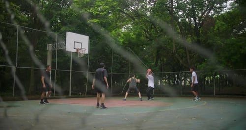 Gruppe junger Männer, die auf einem von Bäumen umgebenen Außenplatz Basketball spielen, ein Spieler erzielte als