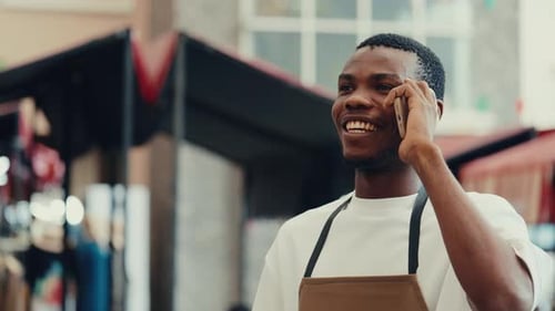 Portrait of African American Male Waiter with Smile Speaking on Phone Against Cafe