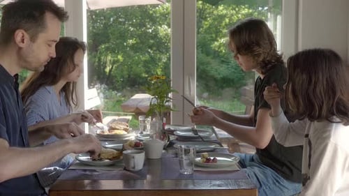 A family is having breakfast at a table by a window overlooking the garden.