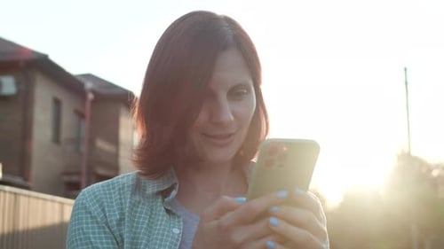 Young Adult Woman Browsing Cellphone Standing Outside In Sunlight