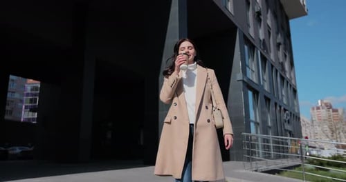 Young Business Woman is Walking on the Street and Drinks Coffee and Using Smartphone