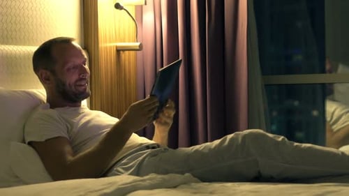 Man relaxes in bed using tablet computer at night