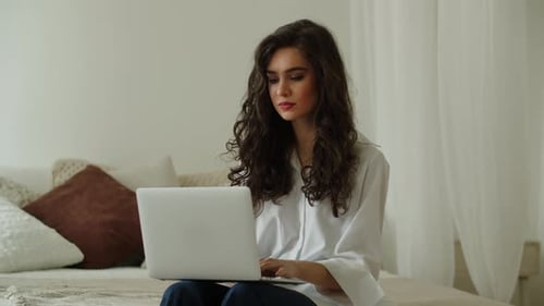 Eine junge Freelancerin arbeitet remote an einem Laptop, während sie zu Hause auf dem Bett sitzt attraktiv