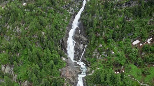 A tall waterfall cascades down a rocky cliff surrounded by dense green forest in summer daylight
