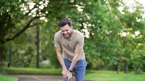 Young adult runner athlete with knee pain in an city park. Man massaging stretching, trauma injury