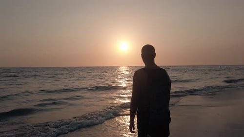 Ein junger Mann am Strand blickt während eines wunderschönen Sonnenuntergangs aufs Meer. Zeitlupenaufnahme.
