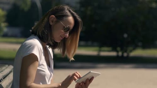 Woman Using Tablet Computer Sitting On Bench In City Park