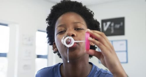 African American child dipping ring wand in pink tub and blowing stream, making bubble in classroom