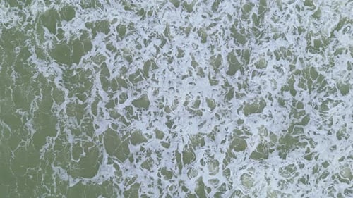 Drone descends to foamy whitewash from behind wave crashing on empty beach, aerial