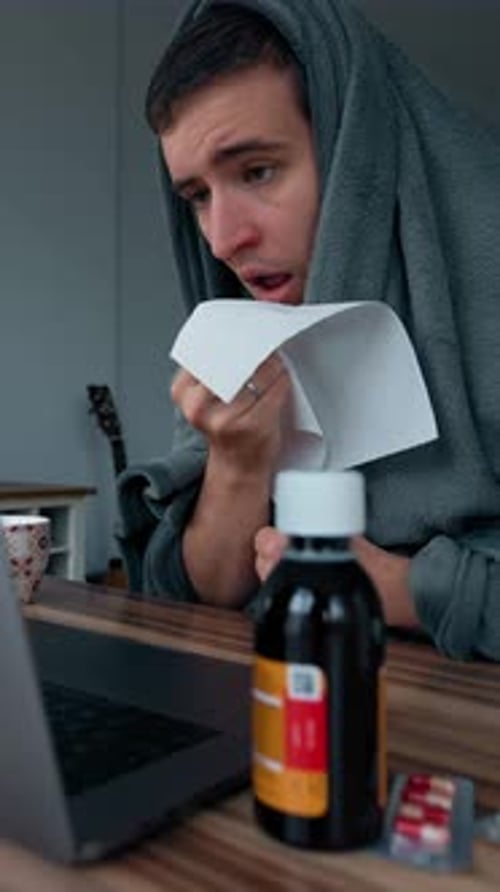 Vertical of Man Wrapped in Blanket Sneezes Covering Mouth with Napkin