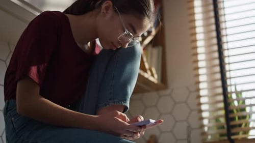 Low angle view of caucasian teenager girl sitting in the kitchen and using mobile phone in silence.