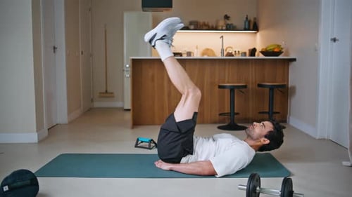 Chico de fitness levantando las piernas acostado sobre una colchoneta deportiva mientras entrena, deportista