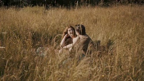 A man and a woman are on a romantic picnic date in a grass field. They laugh together as the camera