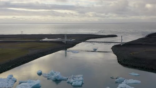 Aerial backwards shot of Icelandic ocean, bridge, river mouth and glacier with icebergs floating on