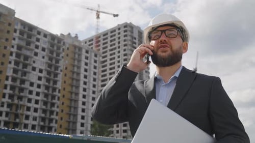 Bearded Engineer Walking with Laptop and Talking on Phone at Construction Site