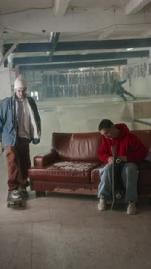Young Guy Fixing Skateboard and Chatting with Buddy at Indoor Skatepark