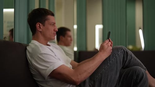 Side View Happy Muscular Handsome Man Sitting on Sofa Using Dating Online Mobile Application Smiling