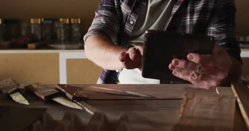 Midsection of caucasian male knife maker in workshop, using tablet and smiling