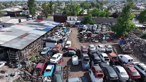 Vordere Drohnenaufnahme eines Schrottplatzes mit Reihen von verschrotteten Autos, einer Lagerstruktur und Wohnhäusern