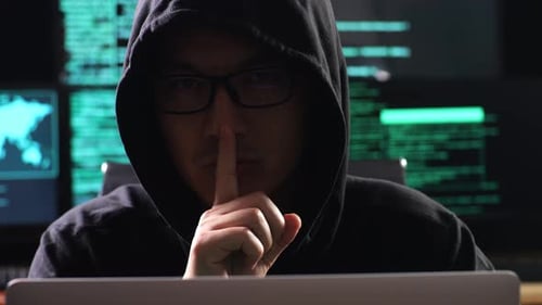 Hacker In A Hood Showing Shh Gesture With Index Finger Near Mouth While Works On A Laptop