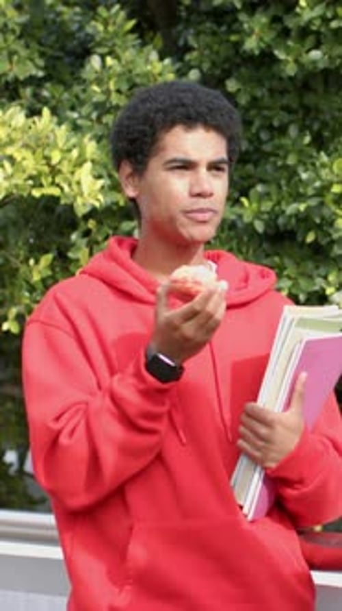 Vertical video: Eating pastry student red hoodie responding to gesturing peer by hedge with folders