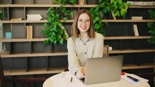 Confident Businesswoman in Glasses and a Shirt Sits at Her Laptop in the Office Looking Directly at