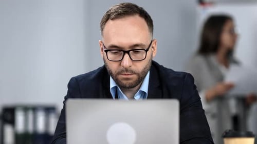 Confident Bearded Man in Eyeglasses Working at Modern Office on Laptop Sitting at Desk Workplace