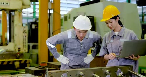 Asian adult male engineer bends over machine frame while Asian adult female technician reviews