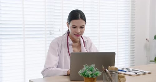 Asian female doctor sits calmly at clinic desk, focused on laptop screen while reviewing patient