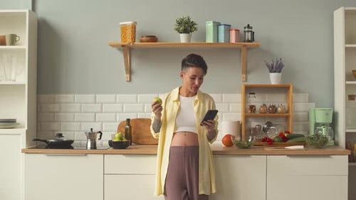 Woman Bites Apple While Checking Cellphone in Kitchen