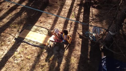 Aerial view of young woman reading book during camping in wild pine forest during sunset time