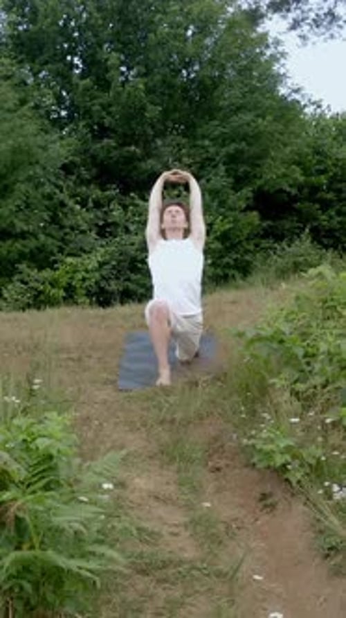 Vertical of Man Practices Yoga Warrior Pose on Mat on Grassy Meadow
