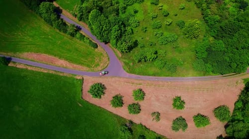 Aerial View of Small Tractor Driving on Winding Rural Road Between Green Fields and Forested Country