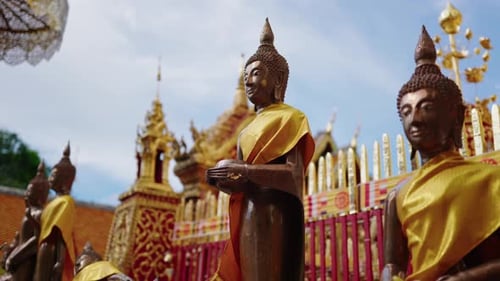 Bronze Buddha statue draped in a golden robe, standing outdoors with an ornate temple structure in t