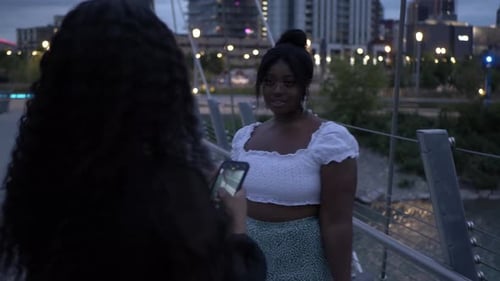 Stylish young woman poses for photo with friend on urban bridge at night