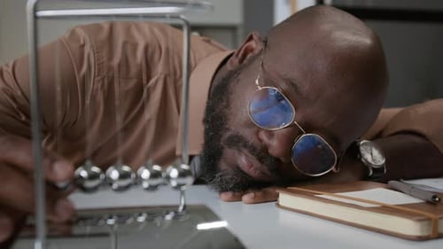 Bored African American Man Looking at Newtons Cradle at Office Desk