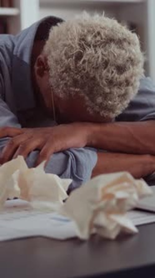 Exhausted African American Man with Blonde Hair Rests His Head on His Arms at His Desk